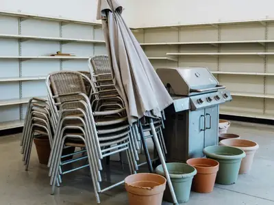 An end-of-season clearance display in a garden centre with stacked patio chairs, a folded patio umbrella, a barbecue and a few garden planters grouped together, half the shelves behind already empty.