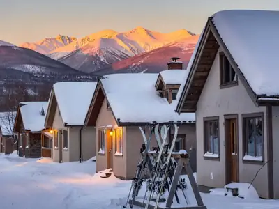 A ski village street at dusk with snow-covered chalet-style lodges, warm lit windows and skis leaning against a rack.