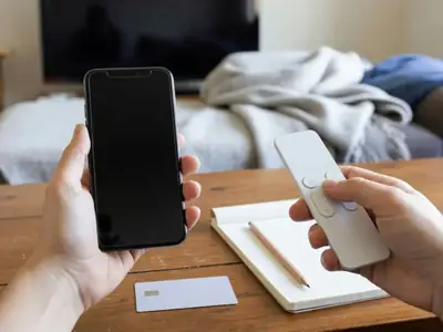 Hands holding a phone and a TV remote beside a blank payment card and notepad in a Canadian living room.