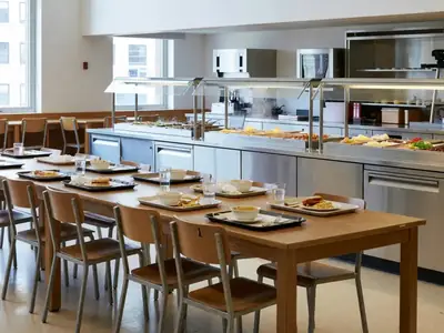 A university dining hall with students eating at long tables and a food serving line.
