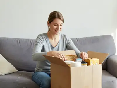 A woman on her sofa unpacking a delivered cardboard box of household supplies and pantry snacks.