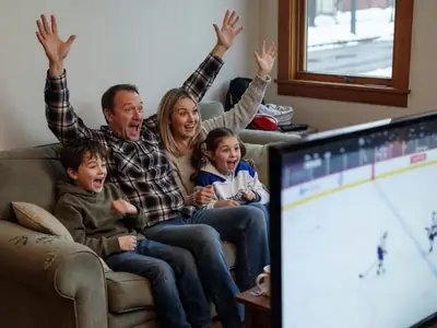 A family watching a hockey game on TV from their sofa, seen from the side, the dad raising his arms as they cheer.