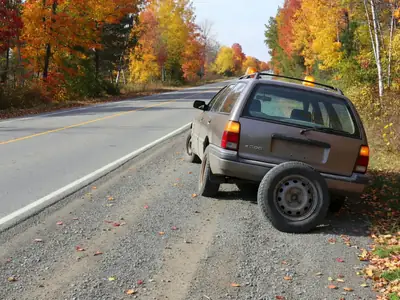 A family car with a flat tire pulled onto the gravel shoulder of a rural highway, hazard lights on.