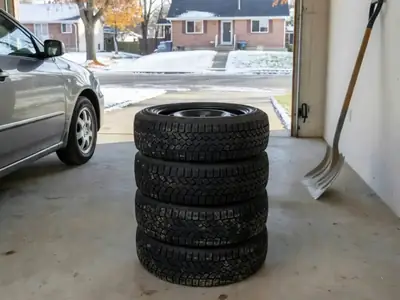 A stack of four winter tires on steel rims in a Canadian home garage beside a car, with the first snow on the driveway outside.
