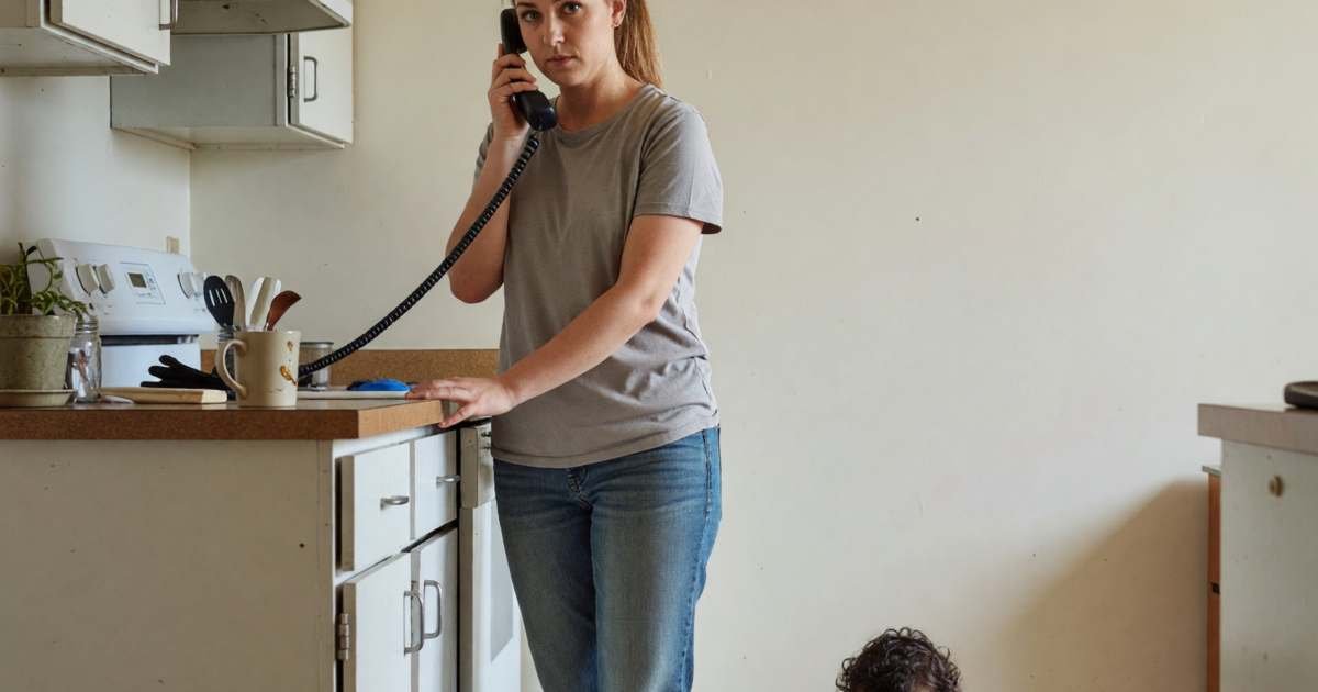 A young mother on the phone at a kitchen counter, calm and determined, with a toddler playing nearby, illustrating setting up a utility payment arrangement before disconnection.