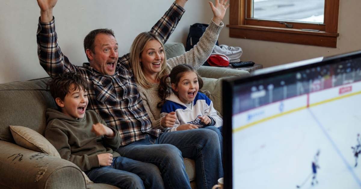 A family watching a hockey game on TV from their sofa, seen from the side, the dad raising his arms as they cheer, illustrating how to watch the NHL in Canada in 2026-27 and what it costs.