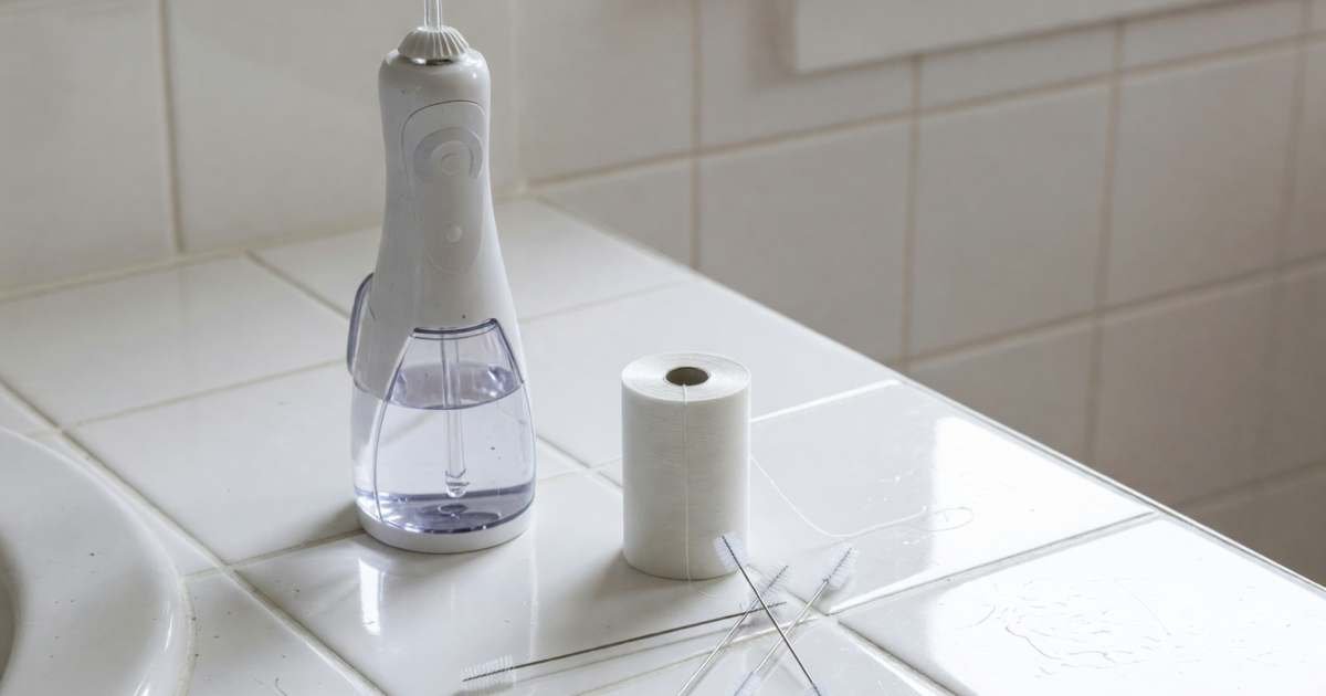 A water flosser, a roll of floss and a few interdental brushes on a bathroom counter, illustrating water flossers versus floss cost per year.